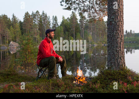 Hiker boiling water in tin kettle over camp fire while camping along lake Stock Photo