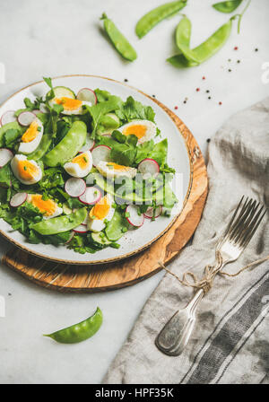Healthy spring green salad with radish, boiled egg, arugula, green pea and mint in white plate on olive tree wood board over grey background, selectiv Stock Photo