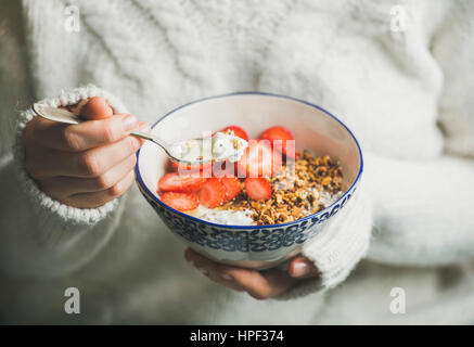 Healthy breakfast greek yogurt, granola and strawberry bowl in hands of woman wearing white loose knitted woolen sweater, selective focus. Clean eatin Stock Photo