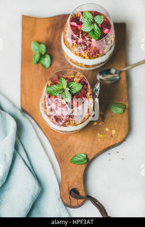 Healthy breakfast. Greek yogurt, granola, blood orange layered parfait in glasses with fresh mint on rustic wooden board over grey marble background. Stock Photo