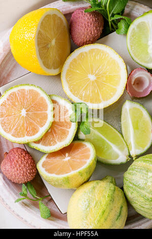 Variety of whole and sliced citrus fruits pink tiger lemon, lemon, lime, mint and exotic lichee on white plate. Close up. Top view. Healthy eating Stock Photo