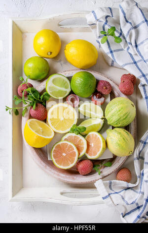 Variety of whole and sliced citrus fruits pink tiger lemon, lemon, lime, mint and exotic lichee on white tray with kitchen towel over white concrete t Stock Photo