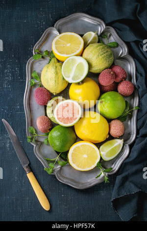 Variety of whole and sliced citrus fruits pink tiger lemon, lemon, lime, mint and exotic lichee on vintage plate with textile and knife over dark blue