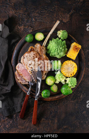 Sliced grilled veal tomahawk steak with vegetables brussels sprouts, romanesco and corn cobs served with cutlery on wooden serving chopping board over Stock Photo