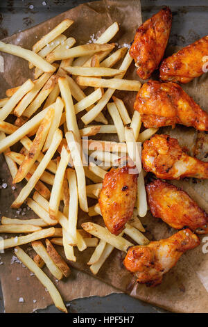 Fast food fried spicy chicken legs, wings and french fries potatoes with salt served on baking paper over old rusty metal background. Top view Stock Photo