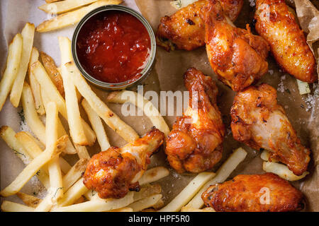 Fast food fried spicy chicken legs, wings and french fries potatoes with salt and ketchup sauce served on baking paper. Top view, close up Stock Photo