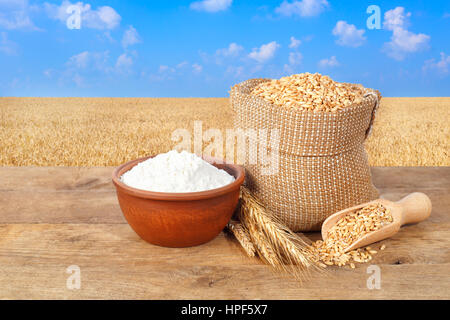 wheat grains in sack. Ears of wheat and wheat grains in bag and flour in bowl on table with field of wheat on the background. Agriculture and harvest  Stock Photo