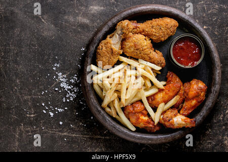 Fast food fried crispy and spicy chicken legs, wings and french fries potatoes with salt and ketchup sauce served in stone plate over dark texture bac Stock Photo