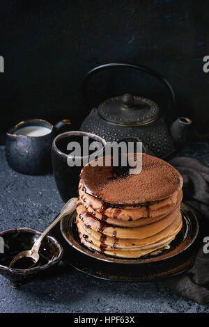 Stack of homemade american ombre chocolate pancakes with carob honey sauce and cocoa powder served on black plate with jug of cream and teapot over bl Stock Photo