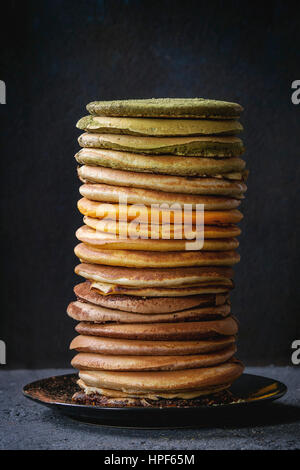 High stack of colorful homemade american ombre chocolate, green tea matcha and turmeric pancakes on plate over black stone texture background. Stock Photo