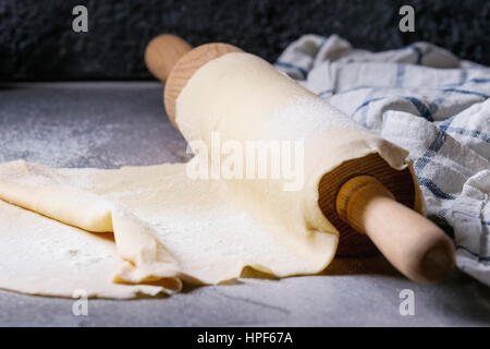Fresh homemade rolled dough for pasta tagliatelle on wood rolling pin with kitchen towel over dark gray table background. Stock Photo