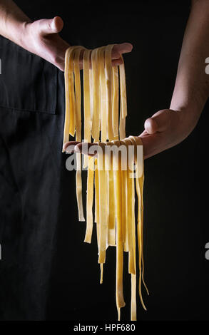 Fresh raw uncooked homemade pasta tagliatelle in man's hands over black apron as background. Stock Photo