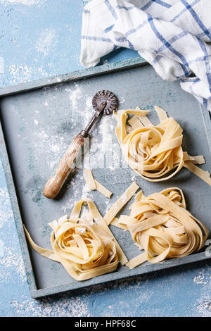 Fresh raw uncooked homemade twisted pasta tagliatelle with vintage pasta cutter in wooden tray with kitchen towel over light blue concrete background. Stock Photo