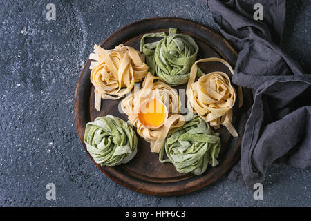 Variety of colored fresh raw uncooked homemade pasta tagliatelle green spinach and traditional yellow with egg yolk on wooden cutting board over dark Stock Photo