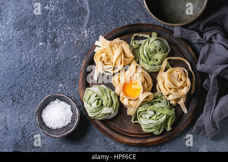 Variety of raw uncooked homemade pasta tagliatelle green spinach and traditional yellow with egg yolk ready to cook with salt on wood cutting board ov Stock Photo