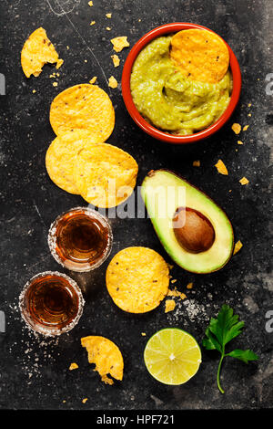 Bowl of mexican nachos chips with homemade fresh guacomole sauce and tequila shots over old  background. Top view. With space for text Stock Photo