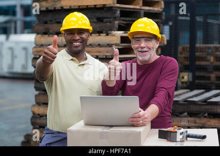 Portrait of two factory workers with laptop showing their thumbs up in drinks production plant