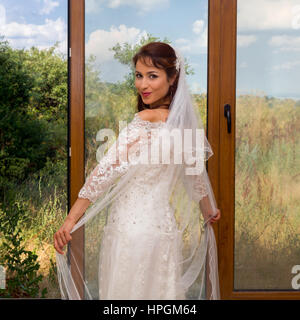 Young bride in traditional wedding dress standing in front of sliding doors or window Stock Photo