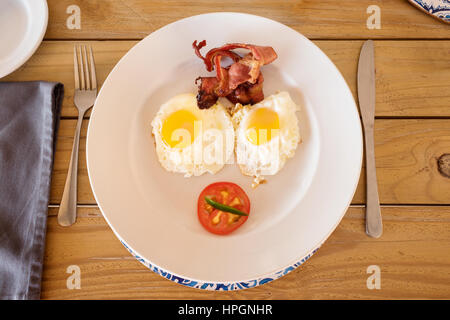 A breakfast plate served up in a camp in Namibia.  On the plate are two fried eggs, sunny side up, some crispy bacon and half a tomato. Stock Photo