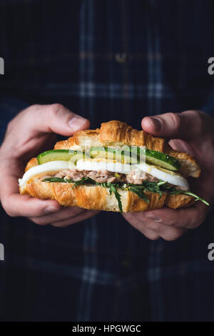 Hands holding tuna croissant sandwich with arugula, egg, tuna salad and cucumber. Closeup view, toned image Stock Photo