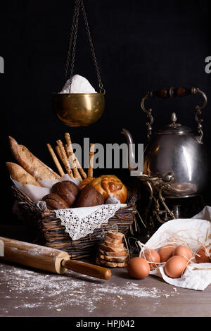 Assortment of freshly baked bread and pastry with eggs, flour and teapot, on rustic wooden table. Stock Photo