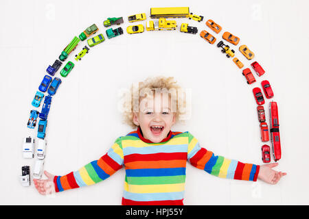 Funny curly toddler boy playing with his model car collection on the floor. Transportation and rescue toys for children. Rainbow toy mess in kids room Stock Photo