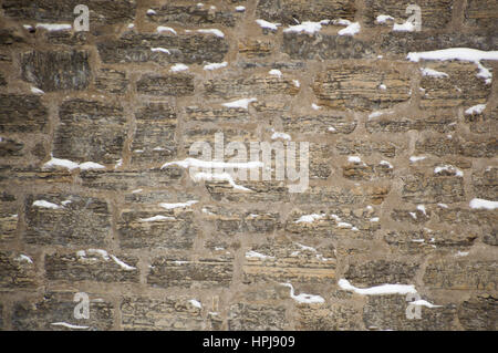 Rugged limestone wall that is uneven and covered with snow in little ledges.  Strata easily seen in the rocks. Rustic and filled with cement mortar. Stock Photo