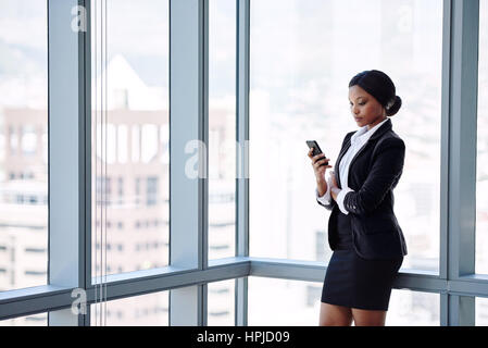 Professional adult female businesswoman busy texting on her cellphone while wearing a white shirt and formal black outfit while standing in front of l Stock Photo