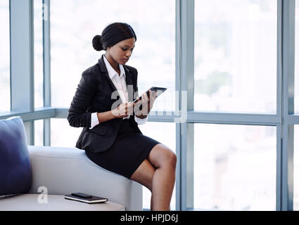 Young black businesswoman sitting on the edge of a sofa in a business lounge while looking at the screen of the digital tablet that she is holding in  Stock Photo