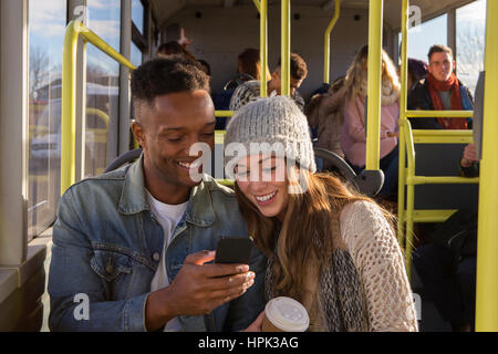 Young couple on a bus. They are both looking at something on a smartphone and smiling. There are people in the background who are also on the bus. Stock Photo