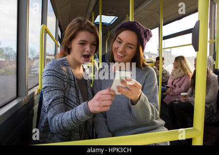 Two young women on a bus. They are both looking at something on a smartphone. There are people on the bus in the background. Stock Photo
