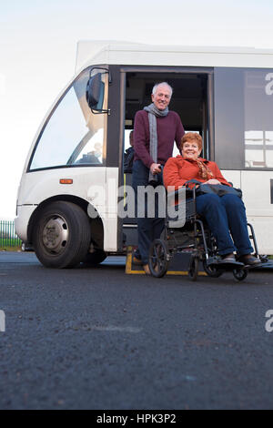 Elderly couple smiling for the camera as they get off the bus. The man is pushing his wheelchair bound wife. Stock Photo