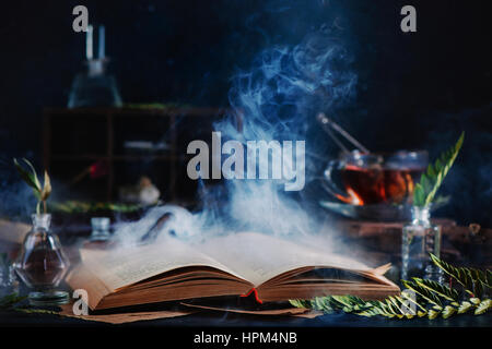 Still life with open spell book, magical smoke, herbs, witchcraft tools and tea Stock Photo