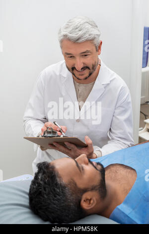Mature doctor writing on clipboard while looking at male patient lying on hospital bed Stock Photo