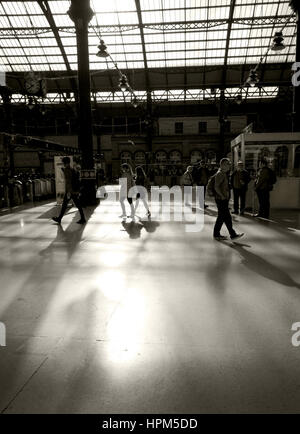 black and white photograph of commuters in train station wlaking to platforms, sun is coming through a glass roof and silhouetting the commuters shape Stock Photo