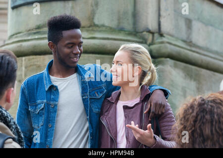 Happy young couple smiling at eachother as they stand outside with friends. They are standing in front of a monument as their friends look up to them. Stock Photo