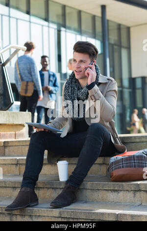 A happy young male smiles as he talks on his mobile phone. He is holding a digital tablet in his other hand. Stock Photo