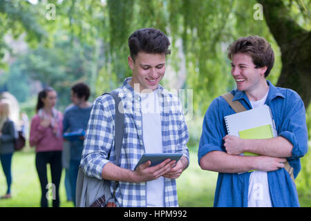 Two happy male students smile as they study together outdoors. They are using a digital tablet and holding books. Stock Photo