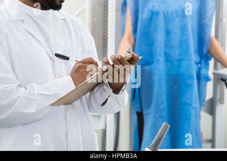 Midsection of male radiologist writing on clipboard with patient in background at hospital Stock Photo