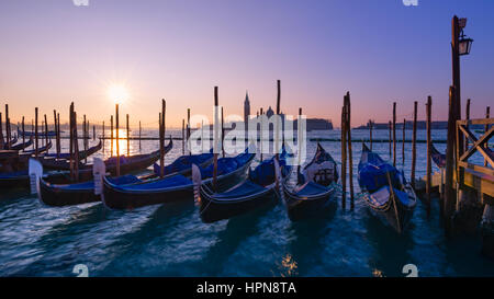 The sun rises above the horizon close to Chiesa di San Giorgio Maggiore. Gondolas can be seen moving in the foreground Stock Photo