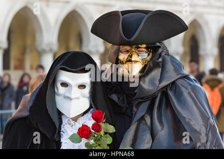 A couple are seen walking alongside the Doge's palace in St Mark's square Venice. they are wearing the traditional Bauta mask and cape, Stock Photo