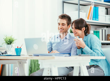 Smiling young couple relaxing at home, they are surfing the web and networking with their laptop, she is holding a cup of coffee Stock Photo