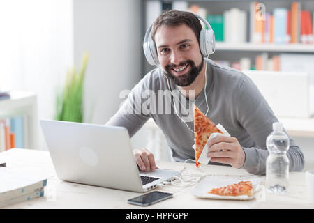 Young handsome man smiling at camera, using a laptop and eating a slice of pizza Stock Photo