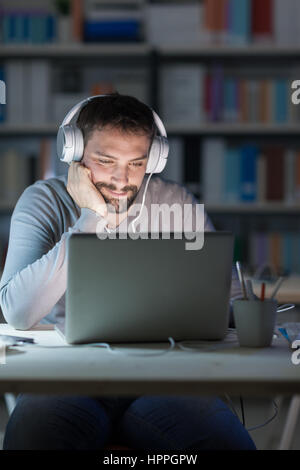 Young smiling man networking and connecting to internet using a laptop late at night, he is sitting at desk and wearing headphones Stock Photo
