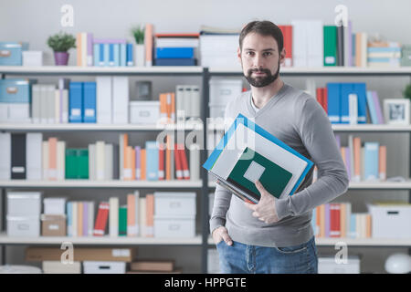 Man holding files and folders and smiling at camera, office shelves with books and files on background Stock Photo
