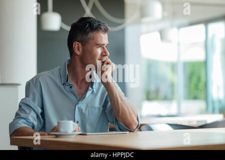 Businessman having a coffee break at the bar, he is thinking with hand on chin, using a digital tablet and looking through a window Stock Photo