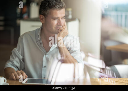 Businessman having a coffee break at the bar, he is thinking with hand on chin, using a digital tablet and looking through a window Stock Photo