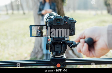 Video shooting at the park: video camera and operator hand on foreground and female model standing in the background Stock Photo