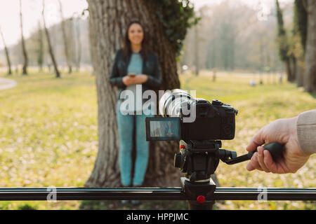 Video shooting at the park: video camera and operator hand on foreground and female model standing in the background Stock Photo