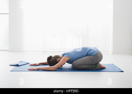 Woman practicing yoga and meditation at home on the floor, she is kneeling on the floor and bending, half tortoise pose Stock Photo